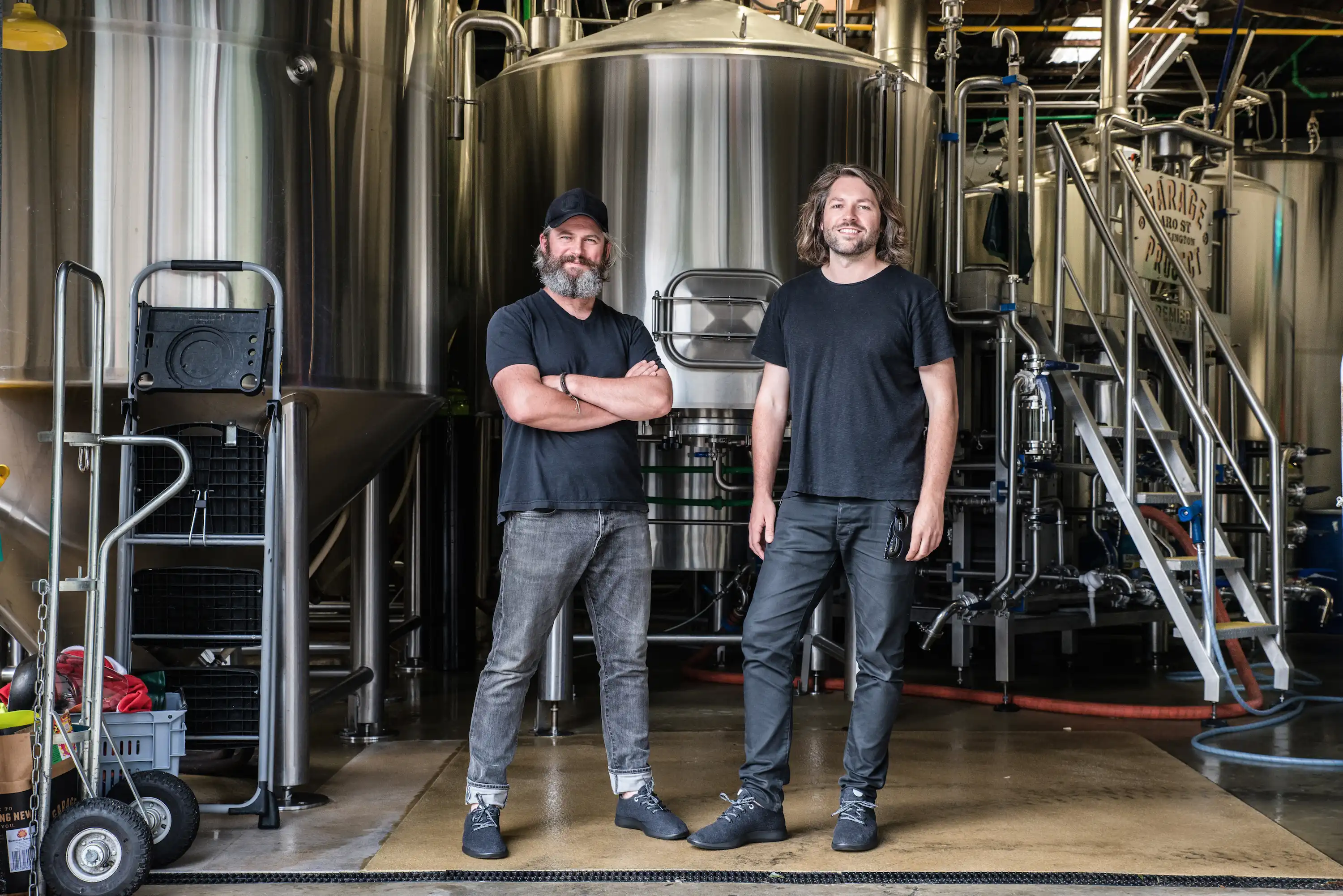 Two men stand in a brewery facility, surrounded by large stainless steel brewing tanks and equipment.