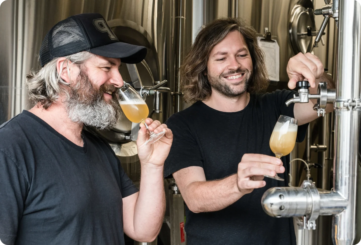 Two men sample beer, one tasting from a glass while the other operates a tap on the stainless steel brewing tanks.