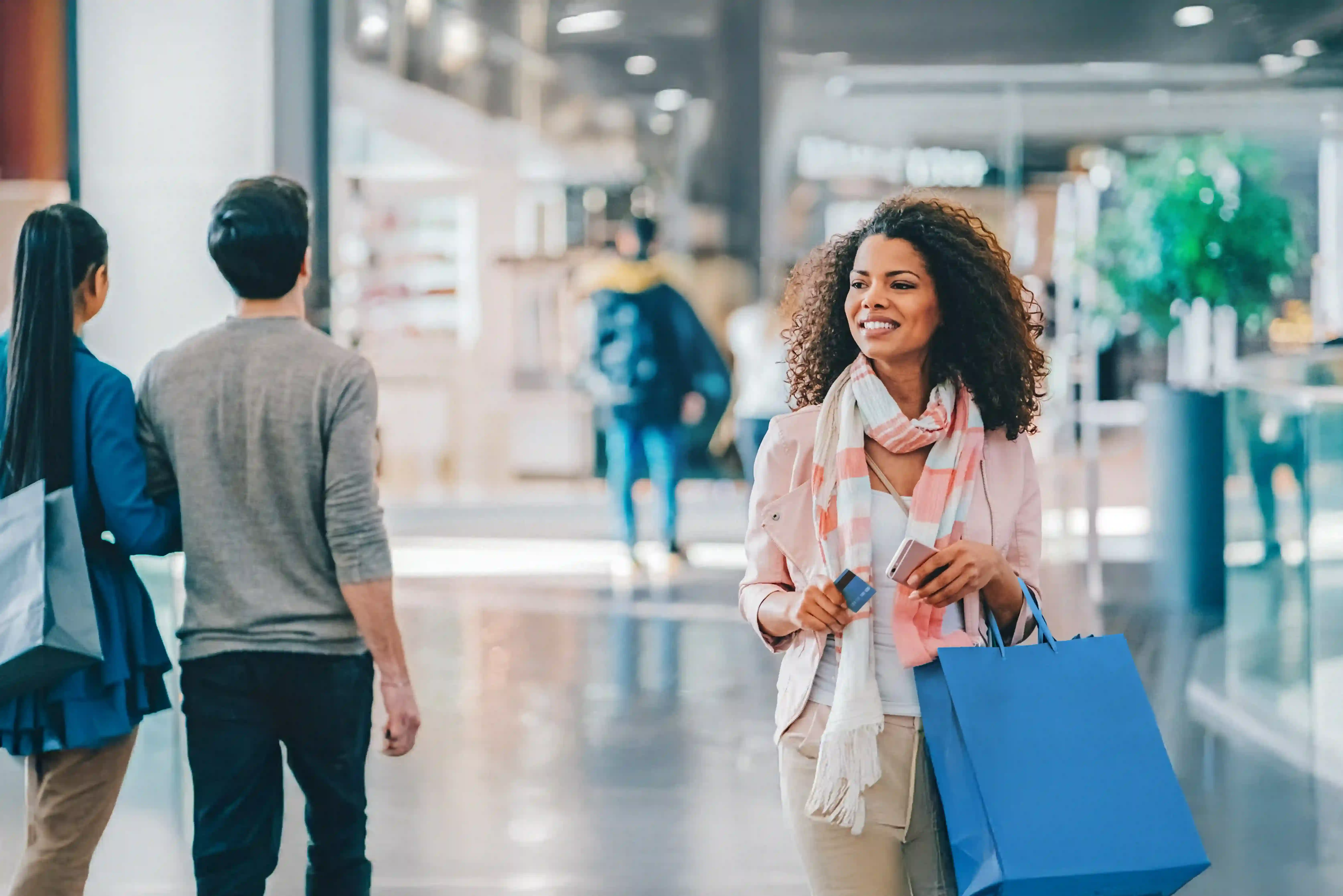 A woman happily shopping in a mall, carrying a blue shopping bag and holding a credit card.